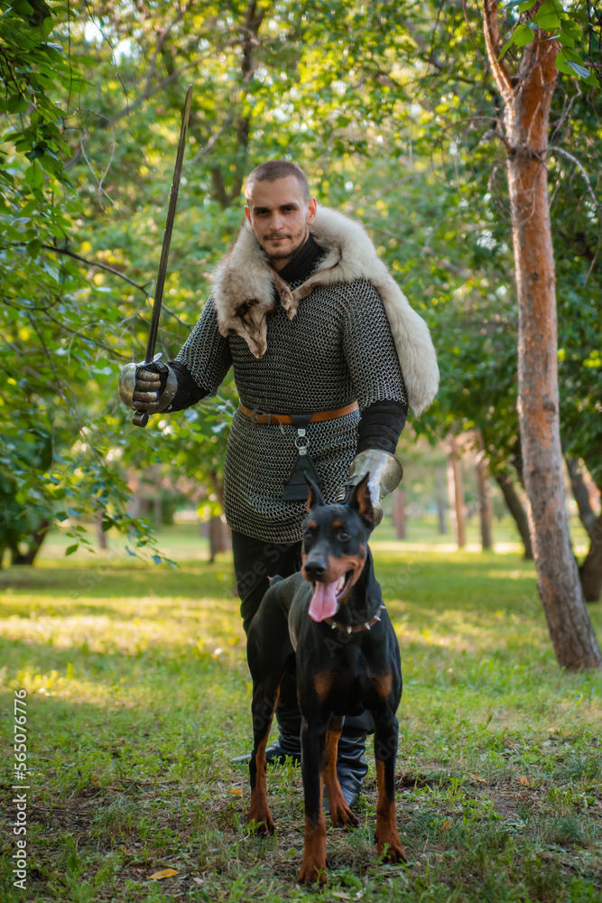 A young man in chain mail with a sword in the woods with a dog. A warrior in armor stands with a Doberman on a chain.