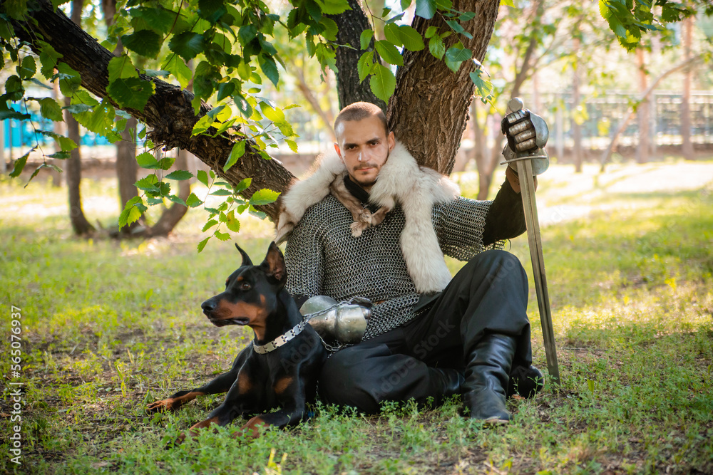 A young man in chain mail with a sword in the forest sits under a tree with a dog. Warrior in armor with a doberman on a chain.