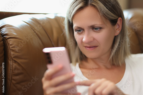 Satisfied woman holds smartphone in her hand and reads messages while sitting on couch.