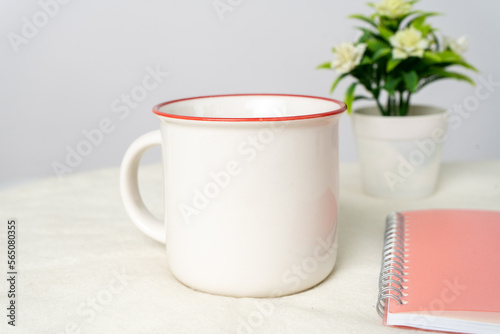 A white blank enamel mug on the top of a white table with some books decorated behind it, minimalistic looks