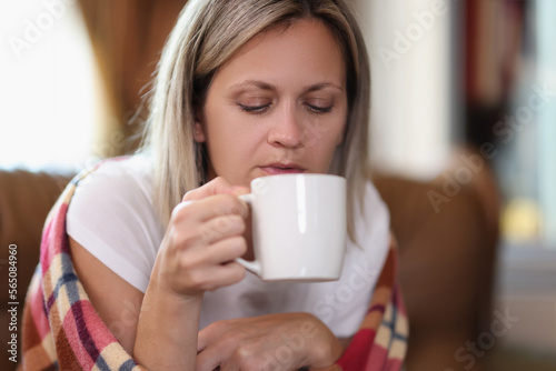 Freezing woman wrapped in warm checkered plaid blanket holds mug and drinks hot beverage at home.