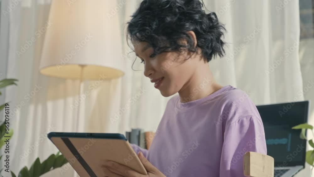 Tilt up shot of happy Asian gen Z girl using digital tablet and then putting on wireless earphones, watch music video and singing while resting at home Tilt up shot of happy Asian gen Z girl using digital tablet and then putting on wireless earphones, watch music video and singing while resting at home
