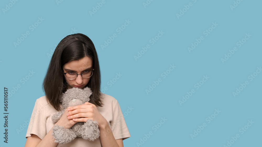 Sad beautiful student girl with glasses, hugging a gray teddy bear on a blue background Sad beautiful student girl with glasses, hugging a gray teddy bear on a blue background