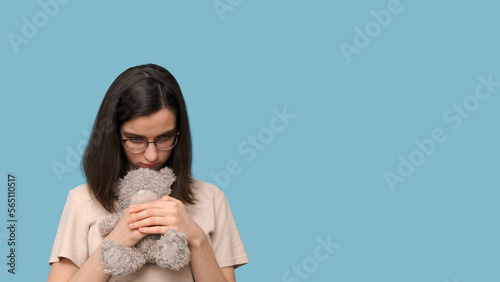 Sad beautiful student girl with glasses, hugging a gray teddy bear on a blue background