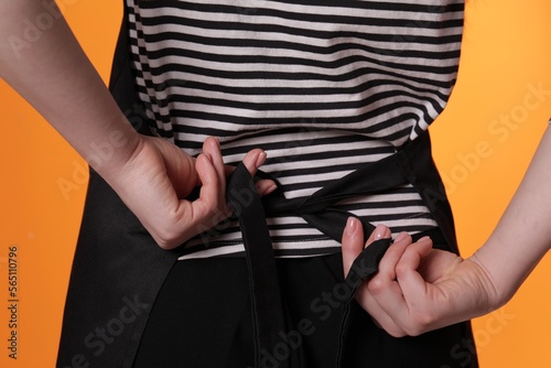Woman putting on black apron against orange background, closeup