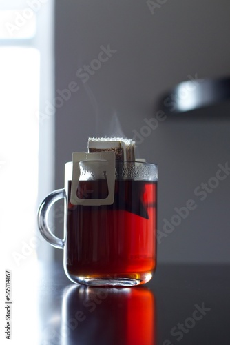 A photograph of a cup of coffee served in a clear glass mug. The coffee is dark in color and appears to be steaming. The mug is resting on a surface and is partially obscured by the steam rising from 