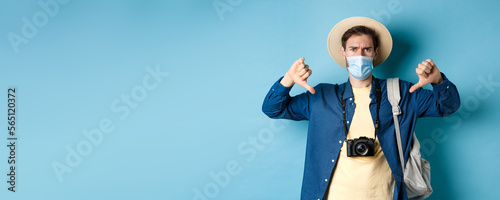 Covid-19 and summer holidays concept. Disappointed tourist in medicla mask frowning, showing thumbs down, negative feedback about travel, standing on blue background photo