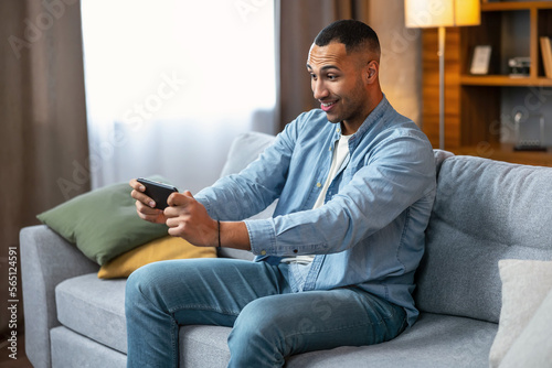 Online gaming concept. Portrait of positive guy playing video games on smartphone at home on sofa