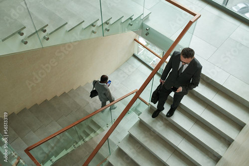 Above shot of two intercultural businessmen in formalwear using mobile phones while walking downstairs inside modern office center