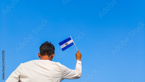 Boy holding El Salvador flag against clear blue sky. Man hand waving Salvadoran flag view from back, copy space