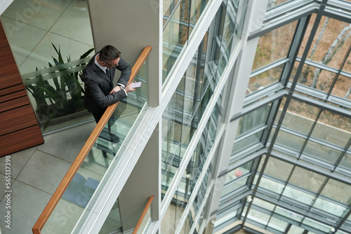 Above shot of young elegant businessman texting in smartphone by railings on upper storey of tall building with large windows