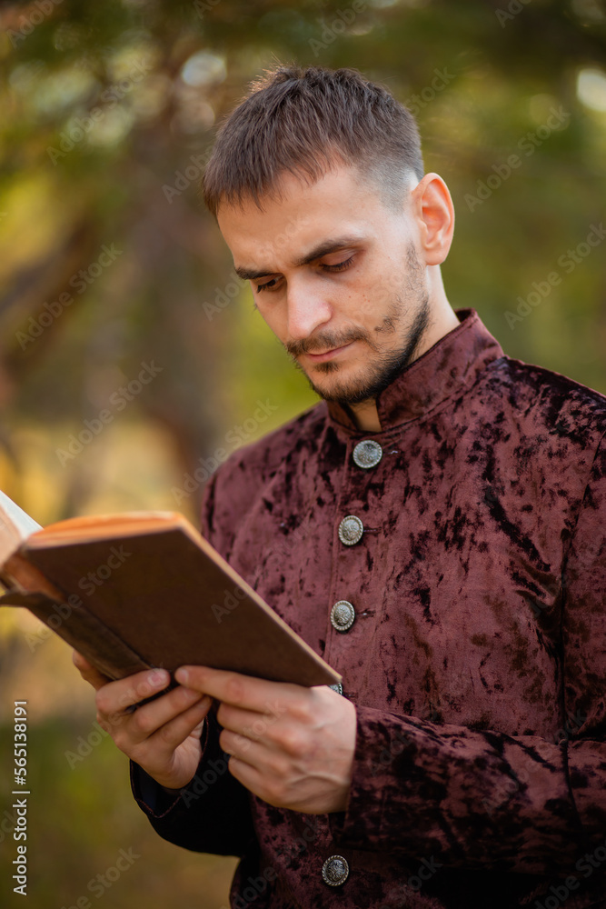 Fototapeta premium A young man in medieval brown clothes stands with an old book in his hand. A guy in a historical costume in nature.
