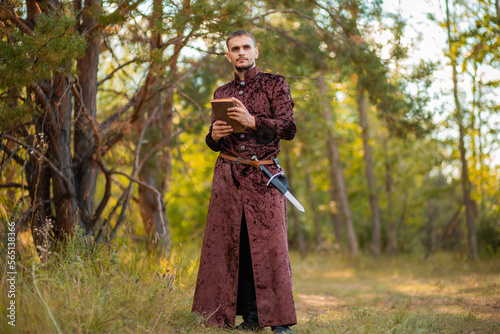 A young man in medieval brown clothes stands with an old book in his hand. A guy in a historical costume in nature.