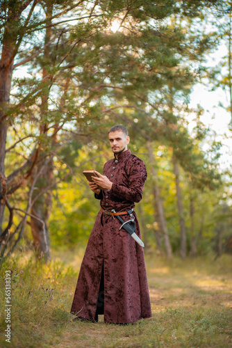 A young man in medieval brown clothes stands with an old book in his hand. A guy in a historical costume in nature. © Вероника Преображенс