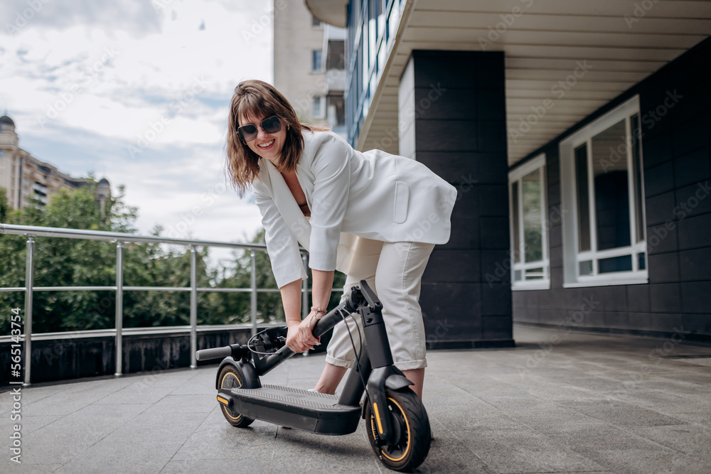 Pretty woman in white suit folding her electro scooter after ride while standing on background of modern building