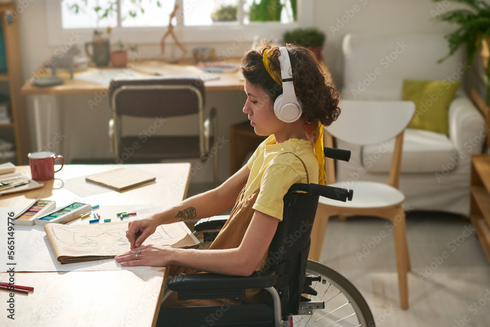 Young woman with disability concentrating on creation of new artwork while sitting in wheelchair and listening to music in headphones Young woman with disability concentrating on creation of new artwork while sitting in wheelchair and listening to music in headphones