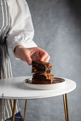 Healthy gluten free chocolate brownie in a plate with nuts and cocoa powder. Homemade, freshly baked delicious brownies in the background and woman's hand takng one. Nutrition concept photo