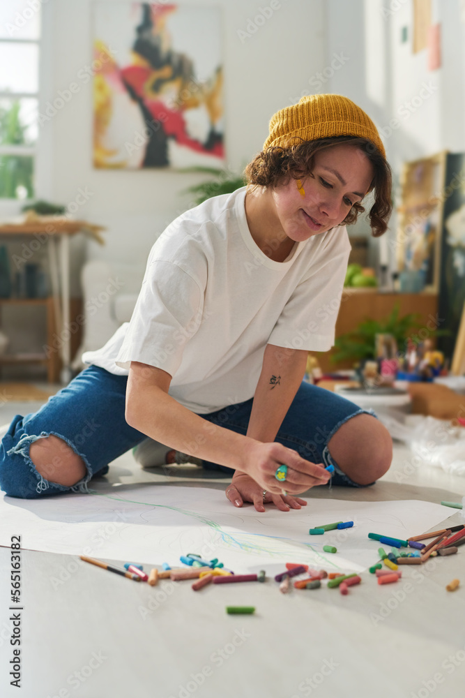 Pretty creative girl in beanie, t-shirt and jeans creating new artwork with crayons on large blank paper sheet while working on the floor Pretty creative girl in beanie, t-shirt and jeans creating new artwork with crayons on large blank paper sheet while working on the floor
