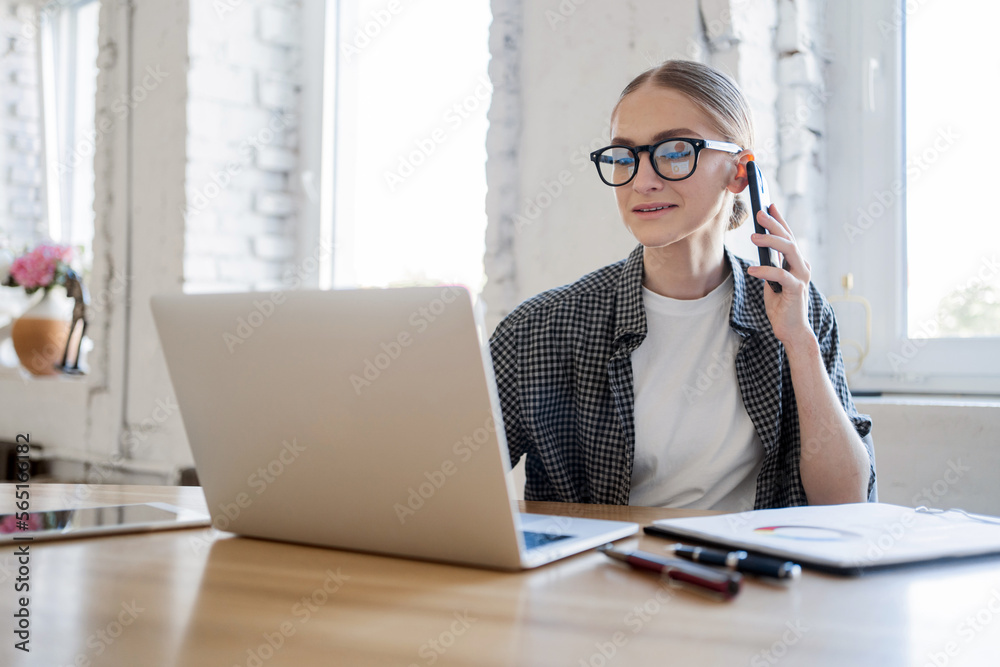 A woman talking on the phone in the office in a shirt, her hair neatly removed, glasses eyes, an online designer working workplace. Coworking space using a laptop computer. A woman talking on the phone in the office in a shirt, her hair neatly removed, glasses eyes, an online designer working workplace. Coworking space using a laptop computer.