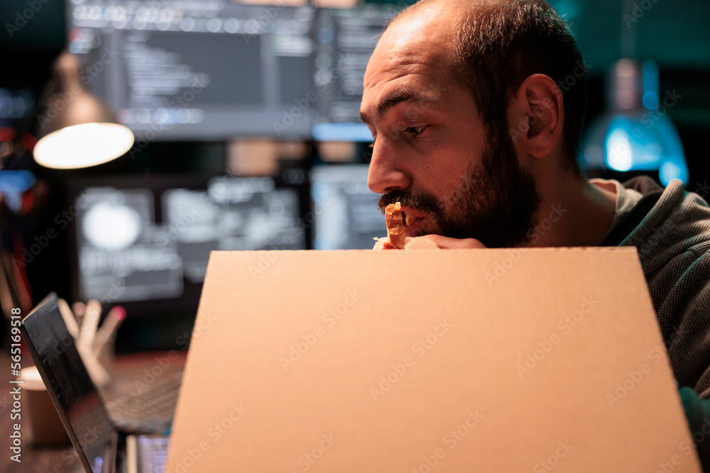 Database coder eating pizza from takeaway delivery, taking bite of food while he works after hours on source code development. Programmer having late dinner with takeout meal in carton box. Database coder eating pizza from takeaway delivery, taking bite of food while he works after hours on source code development. Programmer having late dinner with takeout meal in carton box.