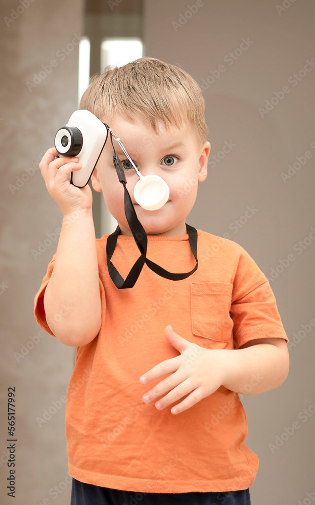 Fototapeta premium People. Handsome funny and emotional caucasian blond little boy in an orange t-shirt with a children's white camera takes pictures in the home interior.
