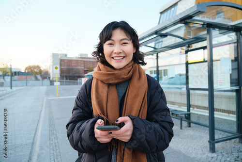 Tela Portrait of Korean woman in winter jacket, standing with smartphone, waiting for