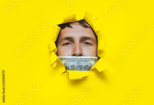 Foto The man's face pokes out through a torn hole in the yellow paper, with a hundred dollar bill and a closed mouth