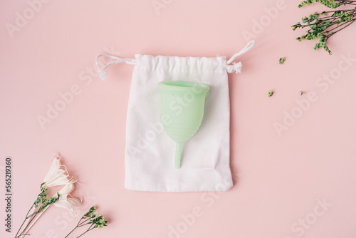 Green menstrual cup on white pouch next to dry flowers on pink background.