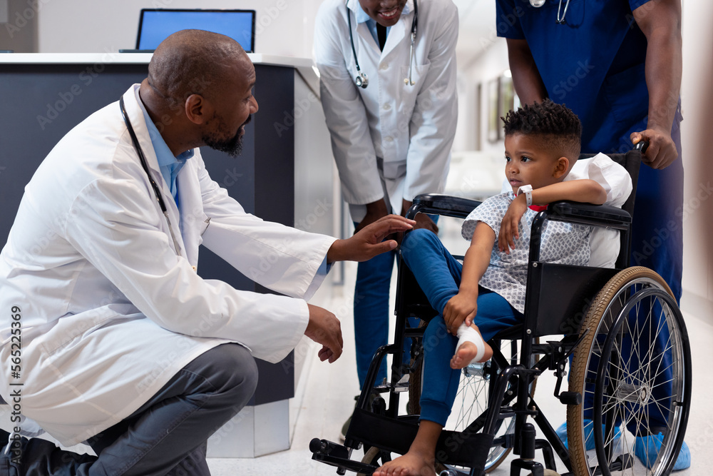 Smiling african american male doctor talking to boy patient in wheelchair in corridor Smiling african american male doctor talking to boy patient in wheelchair in corridor
