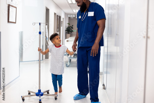 Happy african american male doctor holding hand of boy patient with drip in corridor with copy space