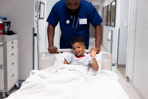 African american male doctor pushing waving boy patient in hospital bed in corridor with copy space