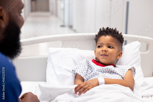 African american male doctor talking to boy patient in hospital bed in corridor with copy space