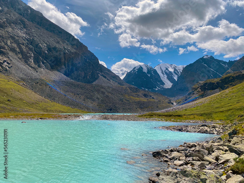 Turquoise mountain lake Karakabak against the background of peaks and stone rocks with glaciers and snow during the day in Altai in the summer among green grass.