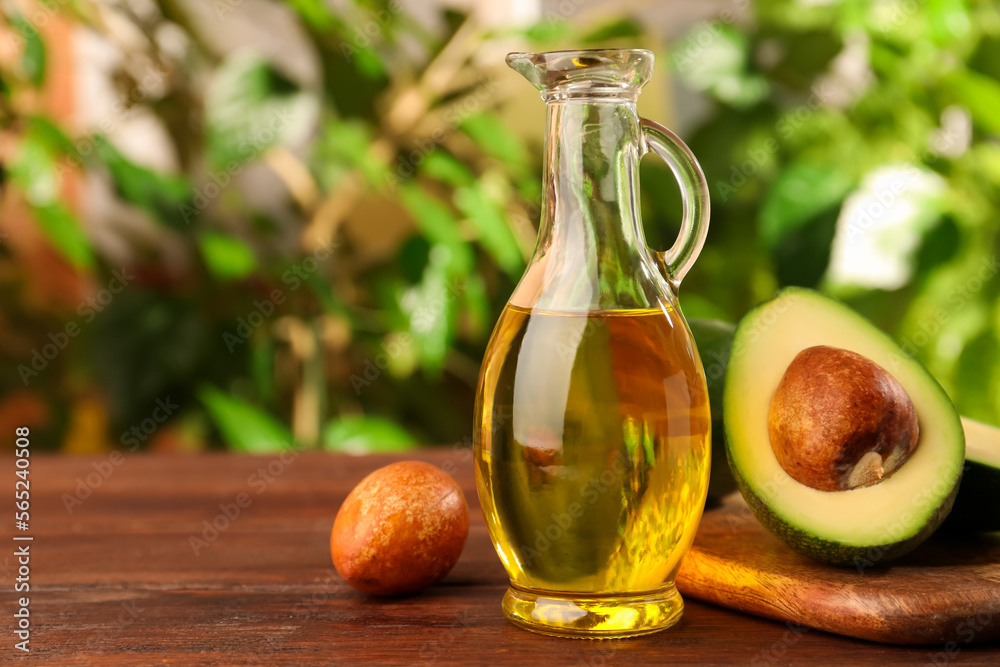 Glass jug of cooking oil and fresh avocados on wooden table against blurred green background, space for text Glass jug of cooking oil and fresh avocados on wooden table against blurred green background, space for text