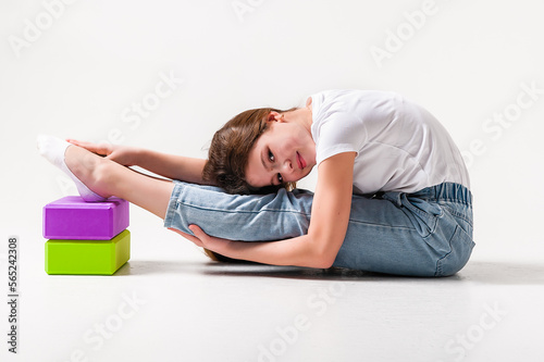 A teenage girl in jeans and a white t-shirt stretches her legs with green and purple styrofoam cubes. White background. The concept of sports, yoga.