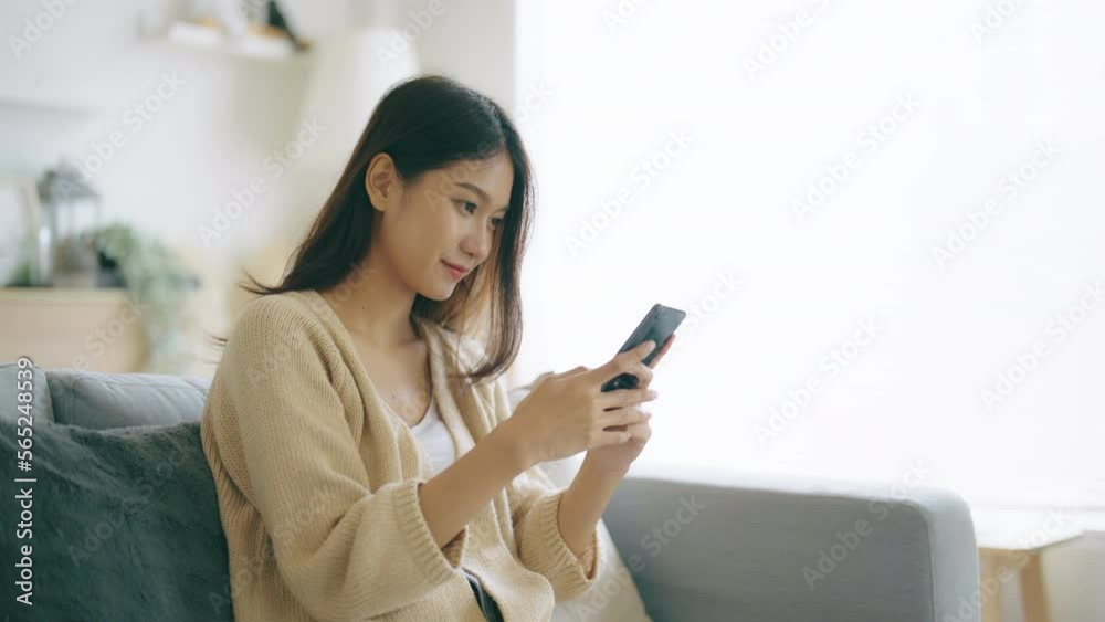 Happy young asian woman relax on comfortable couch at home, typing chat message on smartphone, smiling girl use cell phone chatting, searching information on browser wireless internet, online shopping