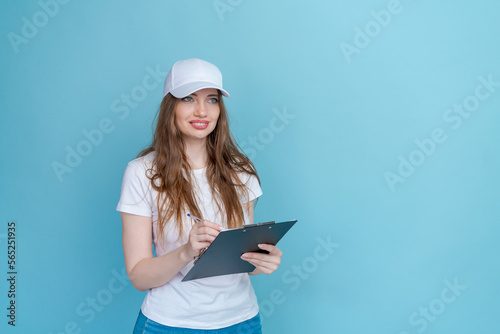 Attractive young caucasian woman in white t-shirt cap blue jeans doing a survey standing against blue copy space studio holding clipboard smiling