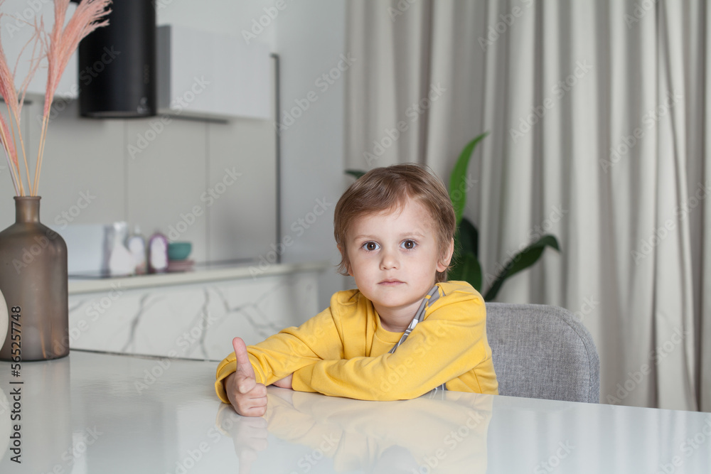 Smiling happy child boy in yellow sweater sitting by the table and showing thumb up on studio kitchen background Smiling happy child boy in yellow sweater sitting by the table and showing thumb up on studio kitchen background