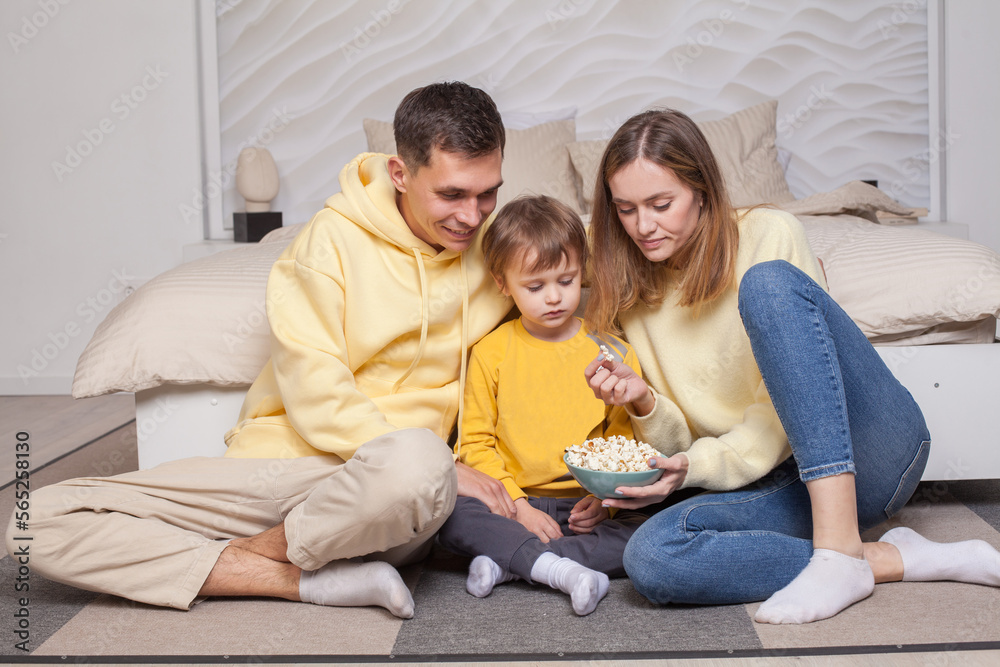 Cute family, parents in yellow clothes with child son sitting by the bed and eating popcorn Cute family, parents in yellow clothes with child son sitting by the bed and eating popcorn