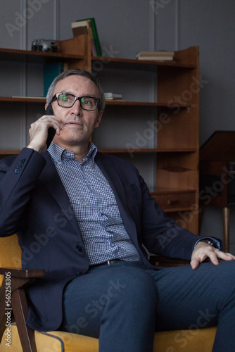 Elderly grey haired businessman in suit and glasses talking about business deals with partners on his mobile phone on grey office wall background