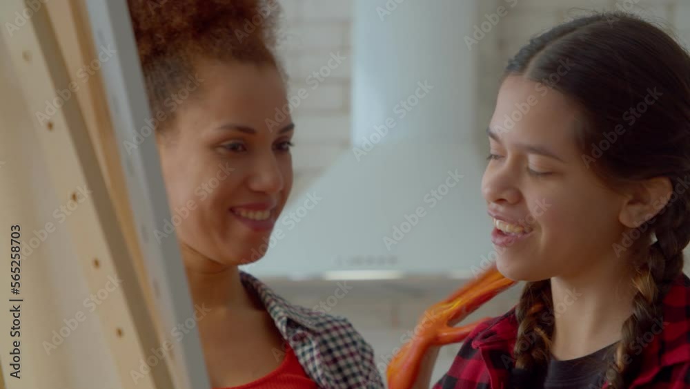 Portrait of positive charming African American mother and adorable multiracial teenager girl with pigtails having fun, creating colorful picture on easel using hand painting technique at home. Portrait of positive charming African American mother and adorable multiracial teenager girl with pigtails having fun, creating colorful picture on easel using hand painting technique at home.
