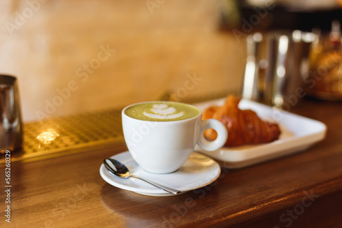 March 13, 2018, Belarus, Minsk. Horizontal photo of a white illy coffee cup with matcha cappucсino and a plate with a croisant on a wooden bar counter.