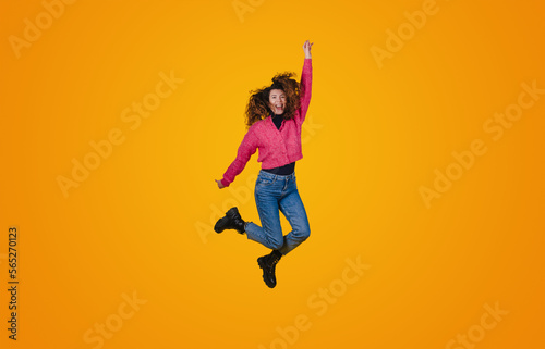 Full length portrait of a cheerful young curly-haired woman celebrating success while jumping isolated over yellow background. Happiness, freedom, power, motion