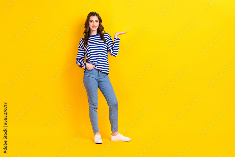 Full size photo of pretty young girl palm hold empty space product promo wear trendy striped garment isolated on yellow color background Full size photo of pretty young girl palm hold empty space product promo wear trendy striped garment isolated on yellow color background