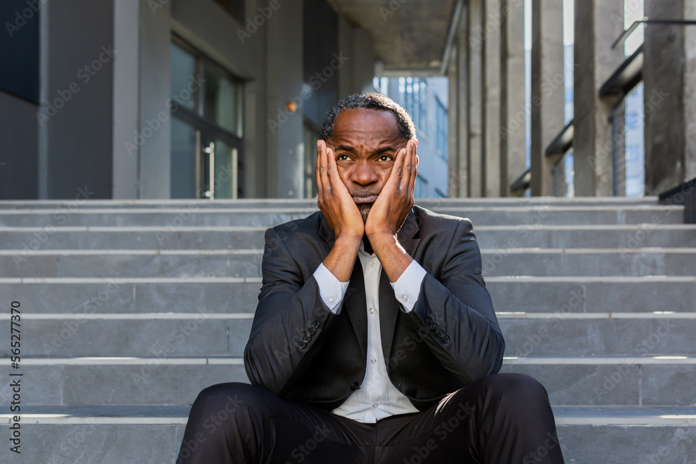 Fototapeta premium Upset african american man sitting on stairs outside office building, businessman bankrupt in despair, mature boss depressed in business suit outdoors.