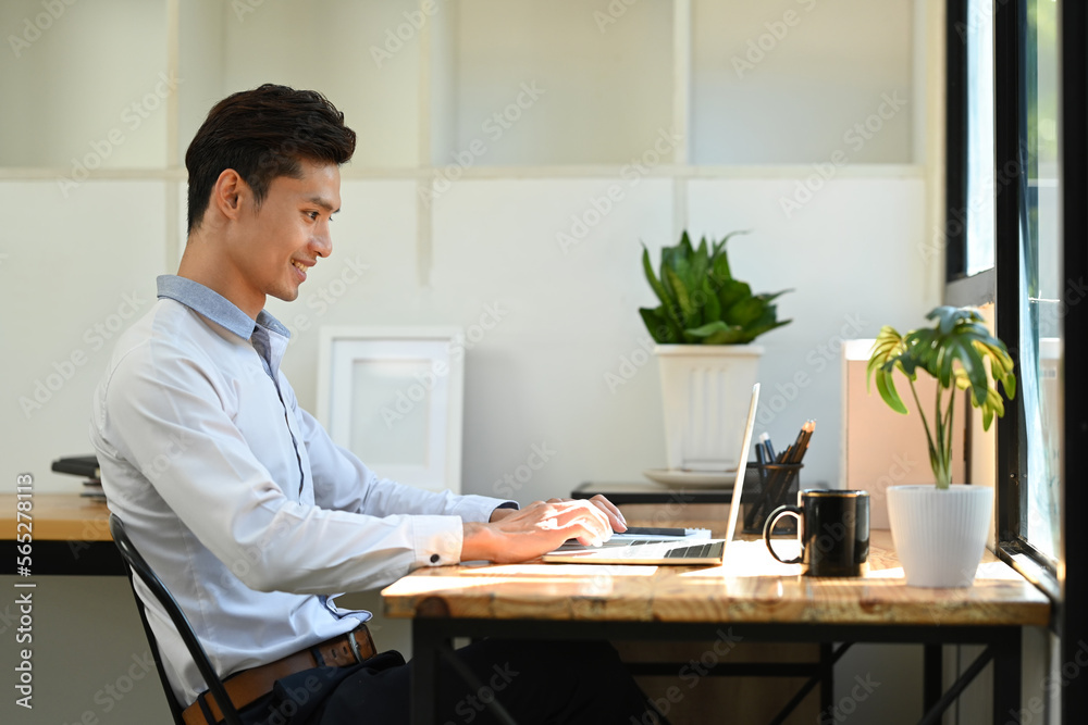 Side view of smiling young asian male investor analyzing financial online data on laptop computer Side view of smiling young asian male investor analyzing financial online data on laptop computer
