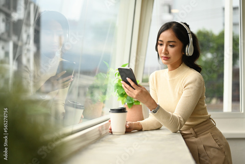 Pretty young woman in wireless headphone chatting with friends on mobile phone while standing near office window