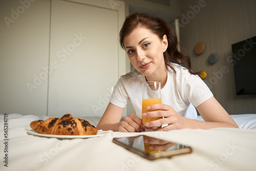 Young woman sits on a bed with croissant and juice