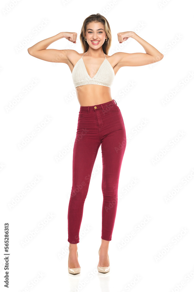 a young woman smiling in burgundy jeans and top, and high heels shoes showng biceps on a white background. Front view a young woman smiling in burgundy jeans and top, and high heels shoes showng biceps on a white background. Front view