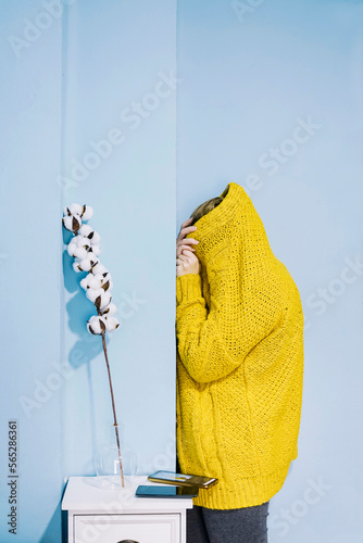 Introvert woman covering herself in yellow woolen coat by blue wall